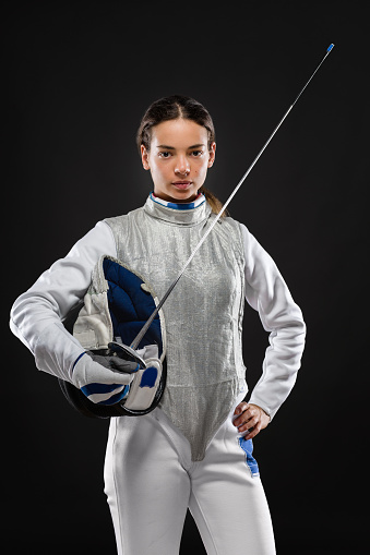 Portrait of Young woman fencer wearing white fencing costume and holding sword and mask. Looking at camera. Black Background