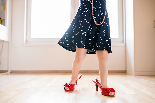 Unrecognizable little girl in dress and red high heels of her mother at home.