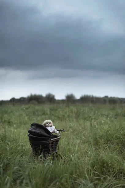 Spooky doll in pram standing in field under dark stormy sky.