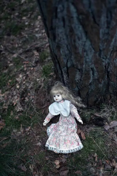 Spooky doll girl on forest ground against tree trunk.