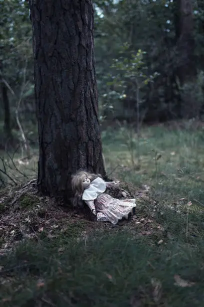 Spooky doll girl on forest ground against tree trunk.