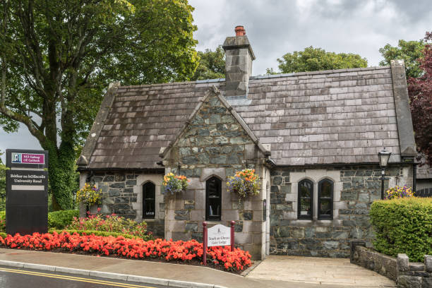 Gate Lodge at main entrance of NUI Galway, Ireland. Galway, Ireland - August 5, 2017: The Gray stone Gate Lodge at main entrance to National University of Ireland Campus stands among green trees and red flowers. NUI sign on the side. galway university stock pictures, royalty-free photos & images