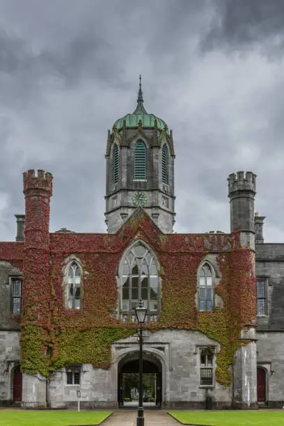 Galway, Ireland - August 5, 2017: Part of historic Quadrangle on National University of Ireland Campus. Closeup of entrance block covered in Ivy with clock tower under dark stormy cloudy sky.