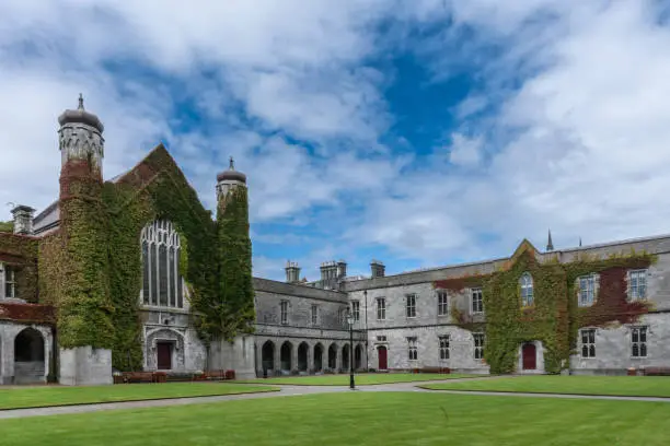 Galway, Ireland - August 5, 2017: Part of historic Quadrangle on National University of Ireland Campus. Quadrangle building covered in Ivy with two towers under blue sky with white clouds. Green lawn.