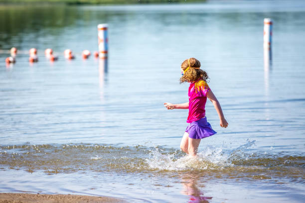 Side view of a young girl splashing water as she walks through the shallow water of a lake on a summer day. The girl has curly red hair and is wearing a pink rash guard swim shirt with purple swim skirt.