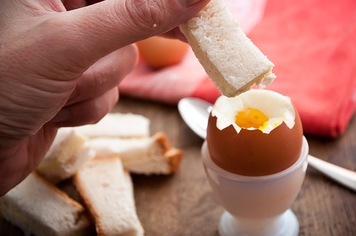 closeup of hand of man eating Soft boiled egg in egg cup and served with toast fingers on wooden table
