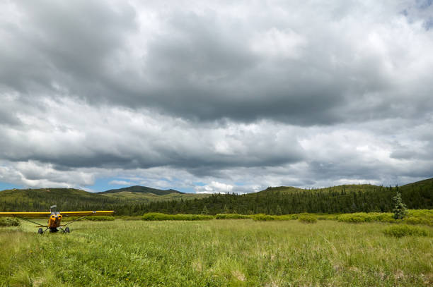 A small, yellow aircraft taking off on a grass, country runway in a scenic location under a cloudy sky.