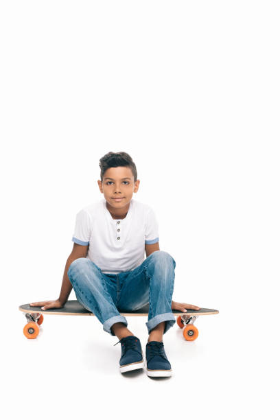 cute african american boy sitting on skateboard and looking at camera isolated on white