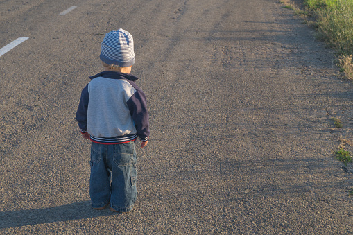 Little boy 1,5 year-old dressed in denim Jeans and sweaters walking along the road. Aspiration or loneliness concept.