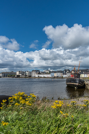 Galway, Ireland - August 5, 2017: Portrait of enormous white cloudscape in blue sky over section of The Long Walk quay. Up front, black ship wreck on dark water in between. Up front, wild yellow flowers and green.