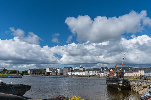 Galway, Ireland - August 5, 2017: Enormous white cloudscape in blue sky over section of The Long Walk quay. Up front, black ship wreck on dark water.