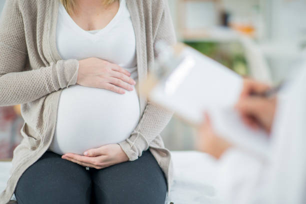 Holding Stomach A pregnant Caucasian woman is indoors in a doctors office. Her female doctor is wearing medical clothing. The woman is holding her stomach which the doctor writes on a clipboard. obstetrician asian stock pictures, royalty-free photos & images