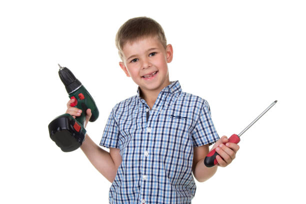 улыбающийся мальчик строитель в клетчатой рубашке держит дрель и отвертку в руках, изолированные на белом фоне - holding screwdriver стоковые фото и изображения