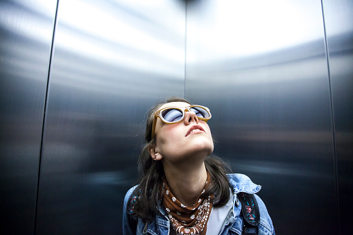 Young woman traveling with elevator and looking up. Wears casual clothes, sunglasses.