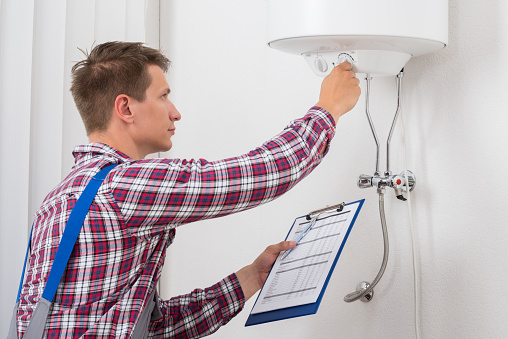 Young Male Plumber Holding Clipboard Adjusting Temperature Of Electric Boiler