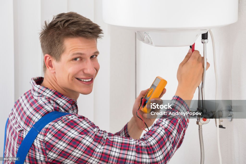 Male Plumber Examining Electric Boiler Smiling Male Plumber Examining Electric Boiler With Multimeter Probe Radiator - Heater Stock Photo