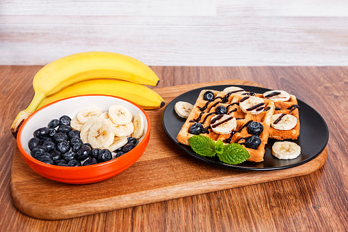 On a wooden cutting board - bunch of banana and a plate with homemade Belgian wafers, poured with chocolate sauce, with sliced banana, blueberries and mint.