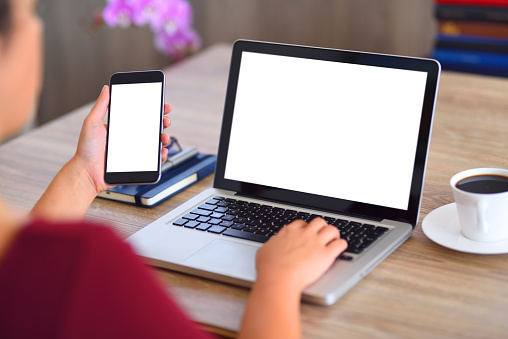 Woman holding a white screen smart phone and  typing on a blank white screen laptop computer on a desk.