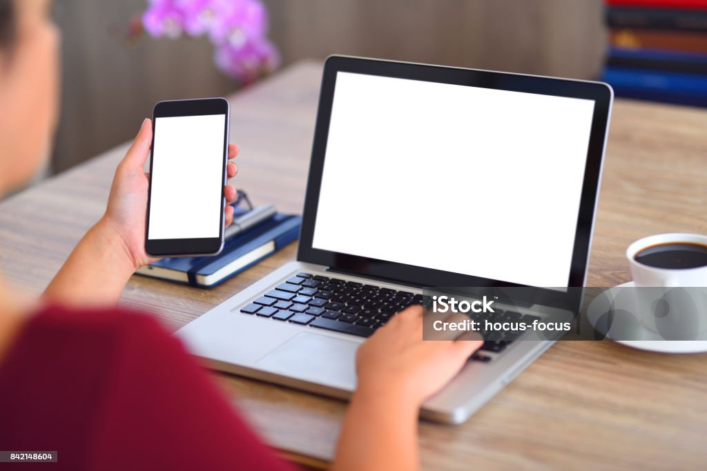 Using laptop computer and smart phone Woman holding a white screen smart phone and  typing on a blank white screen laptop computer on a desk. Laptop Stock Photo