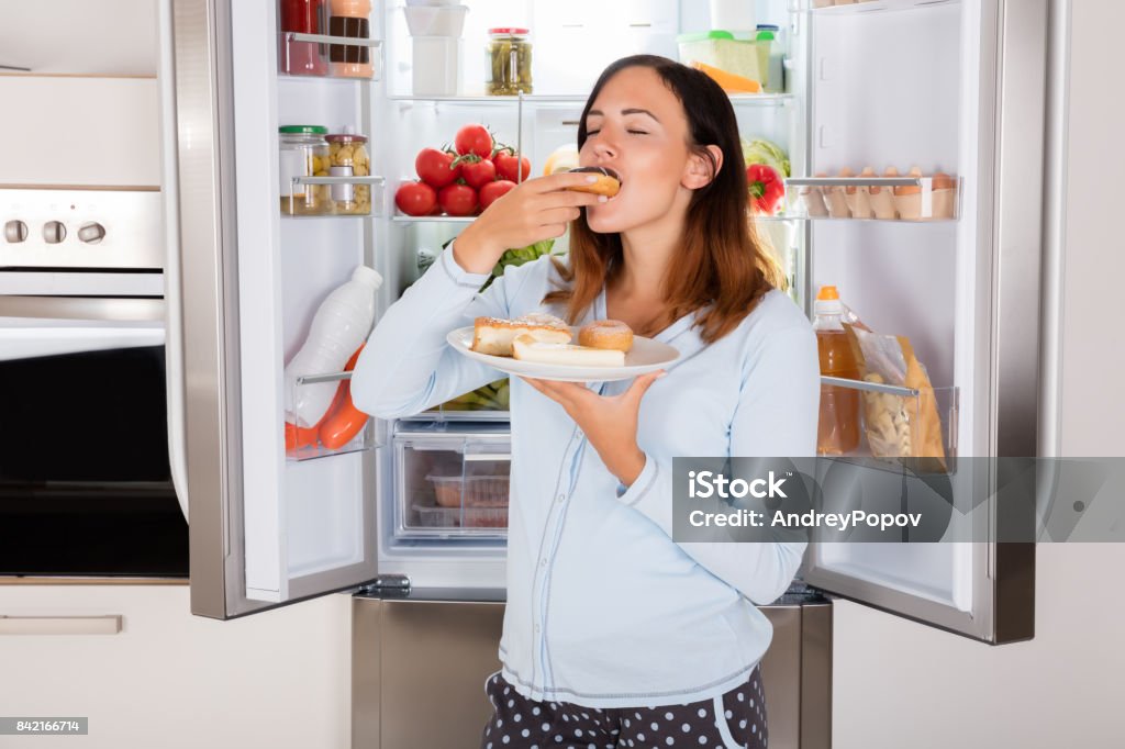 Woman Eating Sweet Food Near Refrigerator Young Woman Enjoy Eating Donut From Plate Near Refrigerator In Kitchen Eating Stock Photo