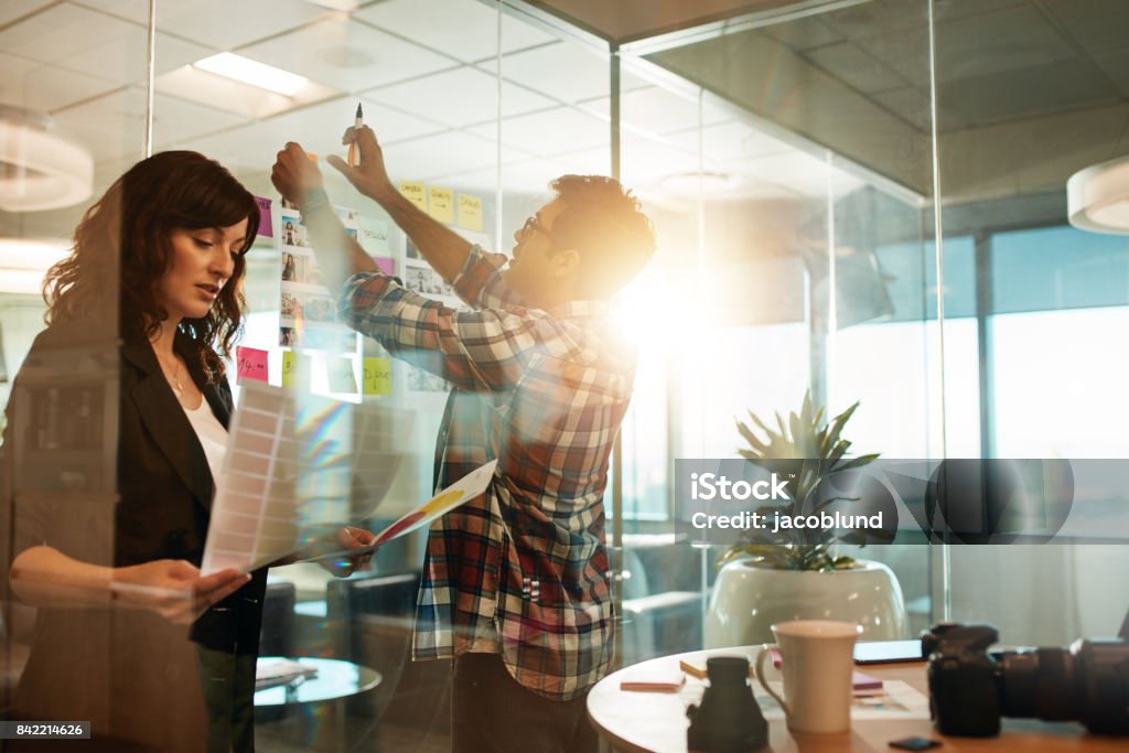 Creative people working on new project Creative people working on new project. Woman looking at photographs and man sticking at contact sheet on glass wall in office. Travel Agency Stock Photo