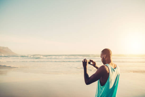 African man photographing on the beach using mobile phone. Young male doing mobile photography at the sea shore.