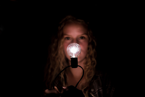 A blurred face of a teenage girl with long hair behind a lit in focus light bulb at night