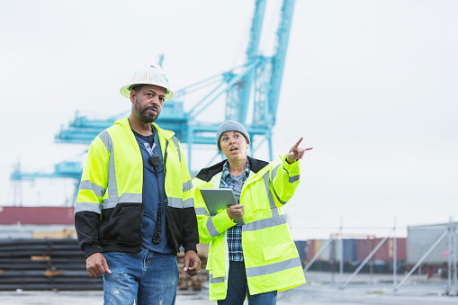 A woman in her 40s and African-American man in his 50s working at a shipping port. They are walking and talking, wearing reflective clothing, with equipment and cargo containers in the background. The woman is carrying a digital tablet.