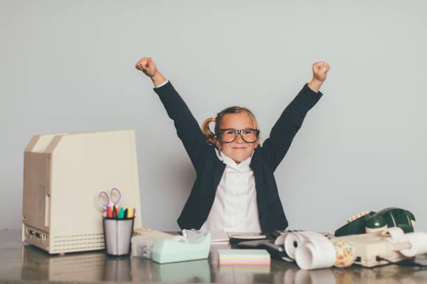 A young businesswoman and girl sits smiling at her office desk happy to help her business make lots of money. She is the business manager in charge of company earnings. She is wearing eyeglasses and a business suit.