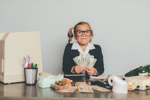 A young businesswoman and girl sits smiling at her office desk happy to help her business make lots of money. She is the business manager in charge of investments. She is wearing eyeglasses and a business suit.