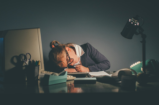 A young businesswoman and girl sleeps late at night at her office desk with her with her head on the desk. She is the financial advisor of her company keeping track of earnings. She is wearing eyeglasses and a business suit.