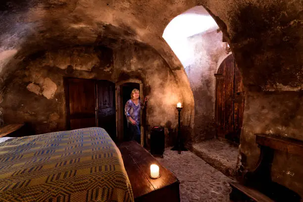 Blond woman in 50s checks into a cave-like medieval guest room with lit candles, massive wooden doors and an arched ceiling in Santo Stefano di Sextantio diffused hotel, Santo Stefano di Sessanio village; Gran Sasso National Park; L'Aquila province, Abruzzo; Italy; Europe