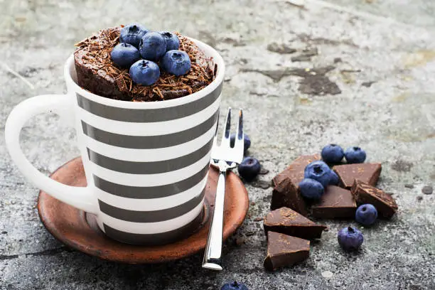 Photo of Healthy breakfast or snack. Chocolate mug cupcake with blueberries and chocolate chips in a gray striped ceramic mug on a gray stone background. Selective focus.