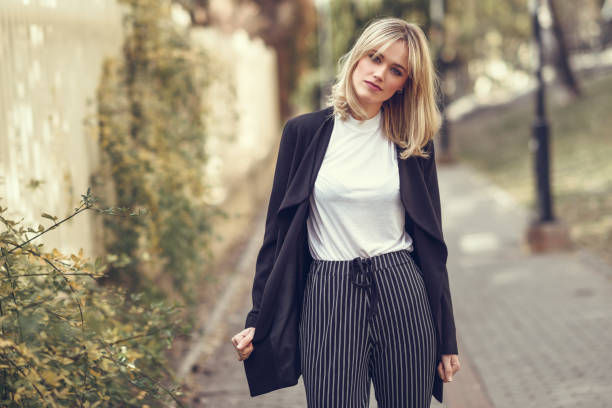Beautiful young blonde woman in urban background Beautiful blonde woman in urban background. Young girl wearing black blazer jacket and striped trousers standing in the street. Pretty female with straight hair hairstyle and blue eyes. blazer jacket photos stock pictures, royalty-free photos & images