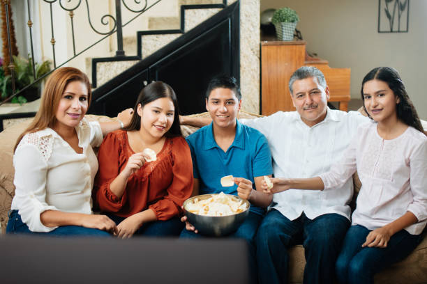 famiglia messicana seduta sul divano e mangiando patatine fritte - family sofa potato chip group of people foto e immagini stock