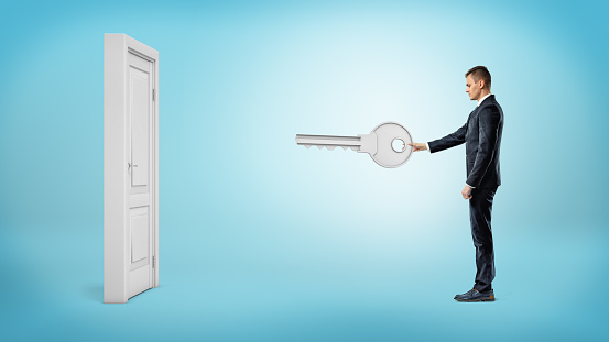 A businessman holds a giant silver key ready to open a white isolated door on blue background. New business opportunities. Door to success. Chance of lifetime.