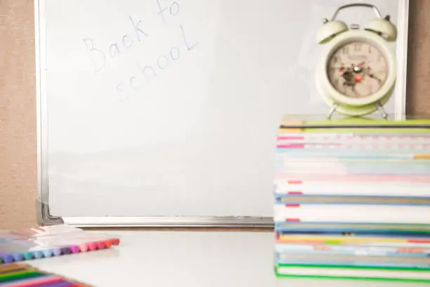 White table with stack of colorful books, alarm clock, small globe, colored pencils and markers with white board in background. Back to school, horizontal view, copy space, kids study time
