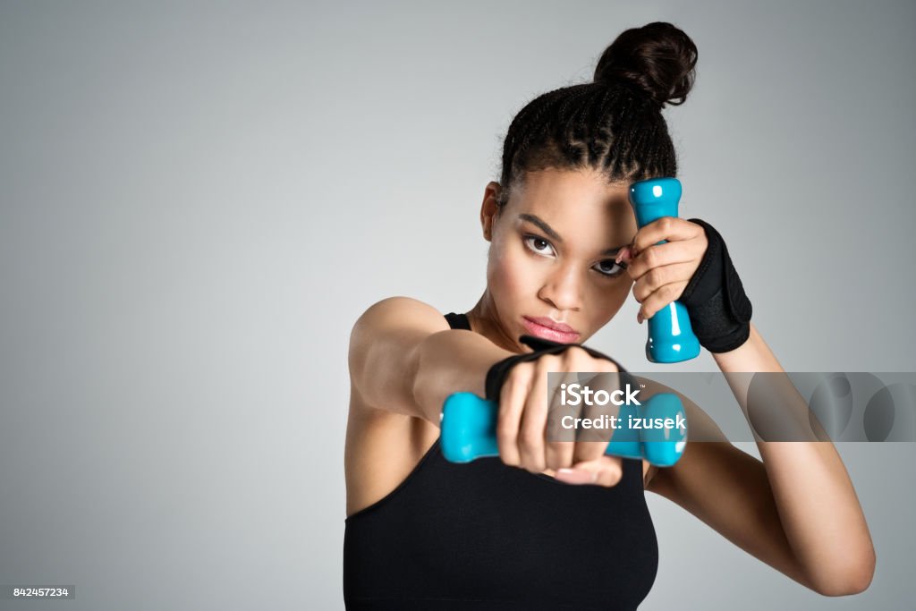 Afro american fit young woman in sports wear holding dumbbell weights Afro american fit young woman in sports wear exercising with hand weights, raising her arms. Studio shot, grey background. 16-17 Years Stock Photo