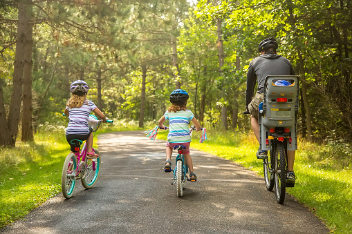 Rear view of a father and his three children (the youngest is in the seat behind him) riding bicycles on a paved path through the woods. Taken on a late summer morning in Minnesota. All four people are wearing helmets.