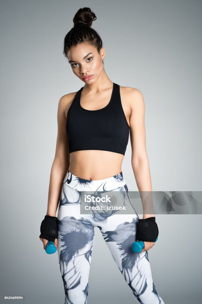 Attractive young woman in sports wear holding dumbbell weights Afro american fit young woman in sports wear exercising with hand weights. Studio shot, grey background. 16-17 Years Stock Photo
