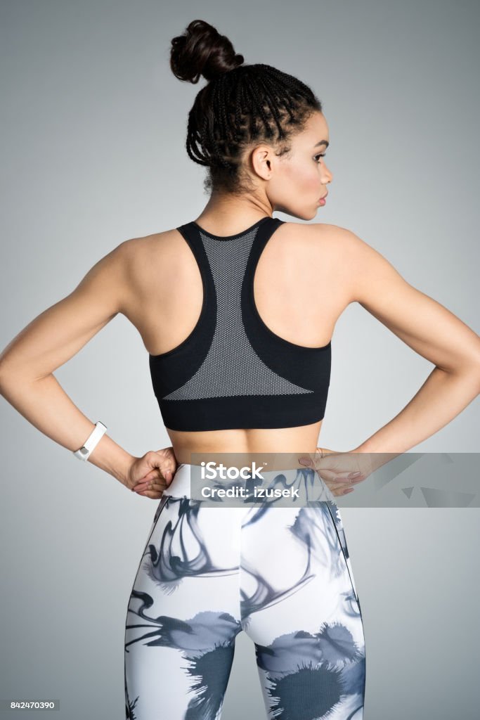 Back view of young woman in sports wear, studio shot Back view of afro amercian young woman in sports wear standing with hands on hips against grey background, studio shot. Back Stock Photo