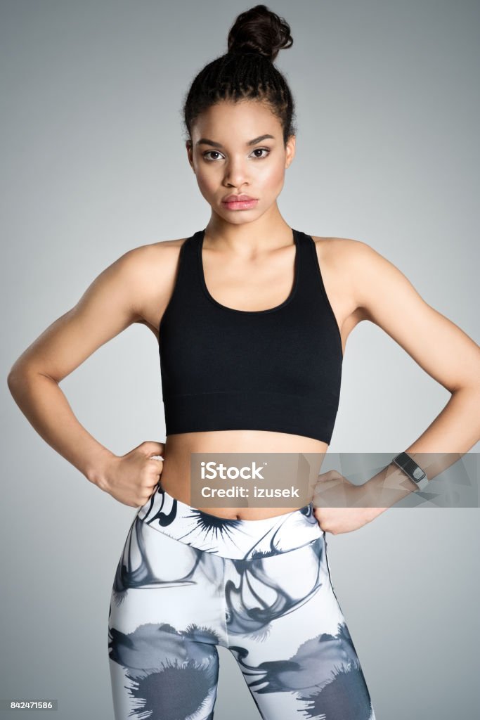 Strong young woman in sports wear, studio shot Attractive afro amercian young woman in sports wear standing with hands on hips against grey background, studio shot. Gray Background Stock Photo
