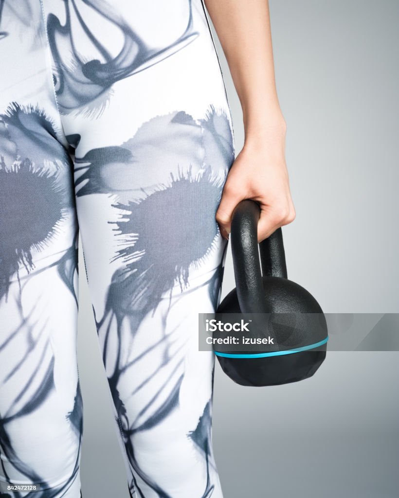 Young woman in sports wear holding kettlebell Part of young woman wearing sports leggings holding kettlebell, unrecognizable person. Studio shot, grey background. Gray Color Stock Photo