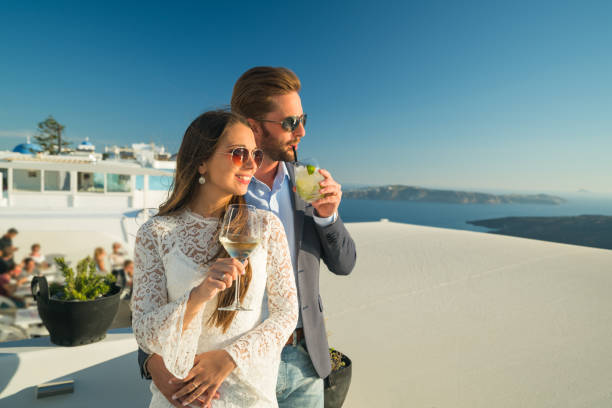 young rich couple with drinks outside restaurant in greece sunset in greece - young couple in evening dress enjoying their drinks greek island of santorini happy couple on vacation in santorini greece stock pictures, royalty-free photos & images