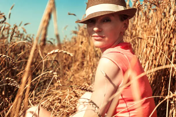The beautiful girl sits in a hat and an undershirt, with a faint smile on a face. Around field of wheat
