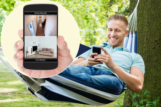 Young Happy Man Sitting On Hammock Looking At Home Security System On Mobilephone