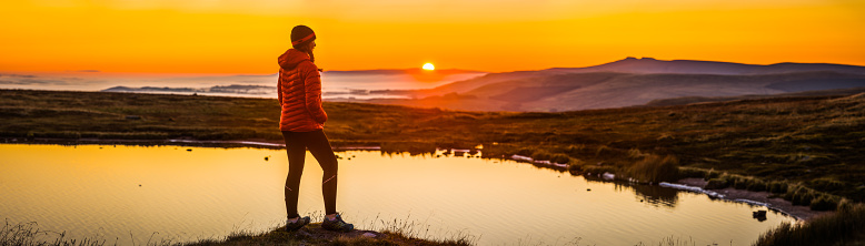 Teenage girl hiker in down jacket and woollen hat looking out over a mountain lake as the sun rises over distant peaks and mist filled valleys.