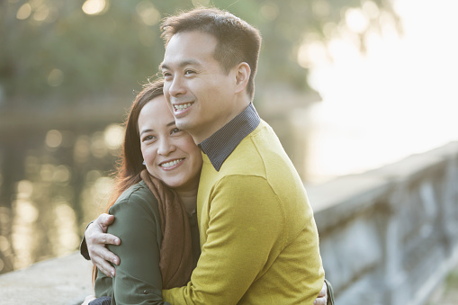 An Asian couple in their 30s embracing, standing in a park by the water. They are smiling, both looking away from the camera in the same direction.