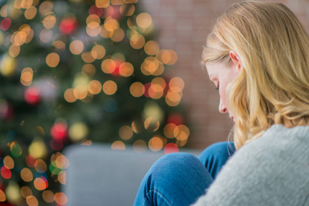 Alone On Christmas A Caucasian woman is indoors in her living room. There is a Christmas tree in the background. The woman is wearing warm clothing. She is sitting on the couch and looking sad because she is alone on Christmas day. sadness stock pictures, royalty-free photos & images