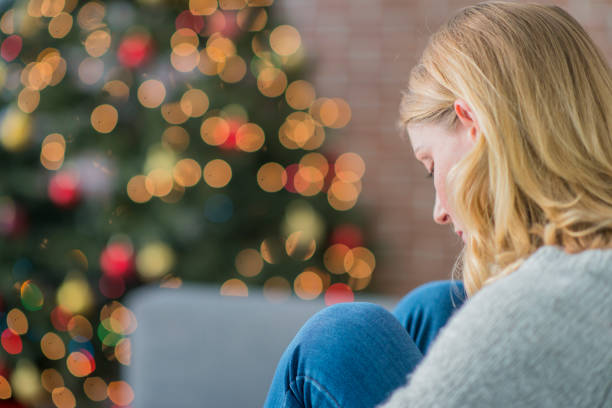 Alone On Christmas A Caucasian woman is indoors in her living room. There is a Christmas tree in the background. The woman is wearing warm clothing. She is sitting on the couch and looking sad because she is alone on Christmas day. unfortnate stock pictures, royalty-free photos & images